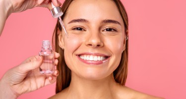 Beautiful woman face, natural clean and fresh glowing skin, applying anti-aging collagen serum. Hands hold serum with active herbal ingredients vitamin C in a dropper, on a pink background.