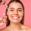 Beautiful woman face, natural clean and fresh glowing skin, applying anti-aging collagen serum. Hands hold serum with active herbal ingredients vitamin C in a dropper, on a pink background.