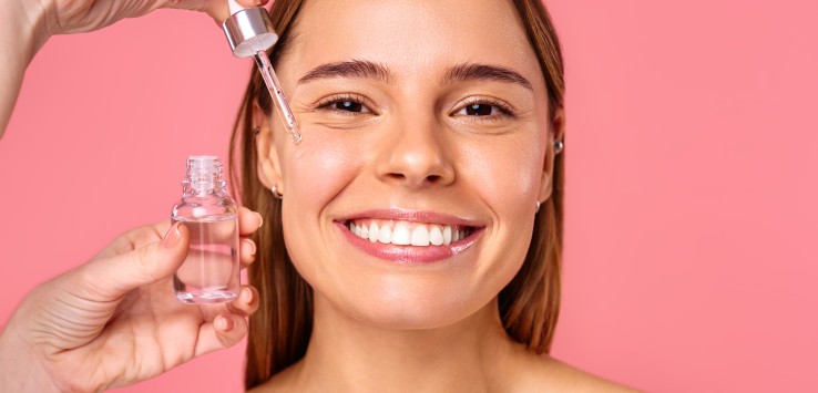 Beautiful woman face, natural clean and fresh glowing skin, applying anti-aging collagen serum. Hands hold serum with active herbal ingredients vitamin C in a dropper, on a pink background.